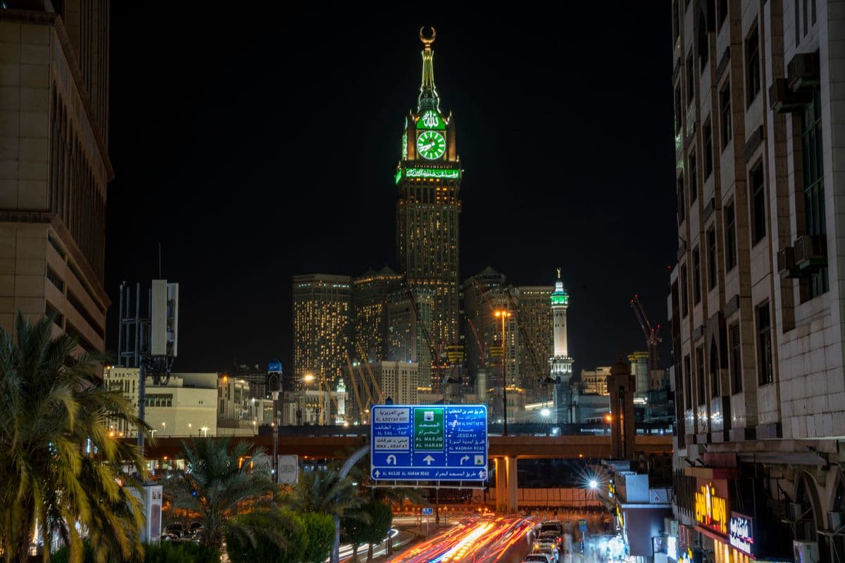 Pemandangan malam kota Mekkah dengan menara jam Abraj Al-Bait yang megah bercahaya, melambangkan kekhusyukan perjalanan spiritual.