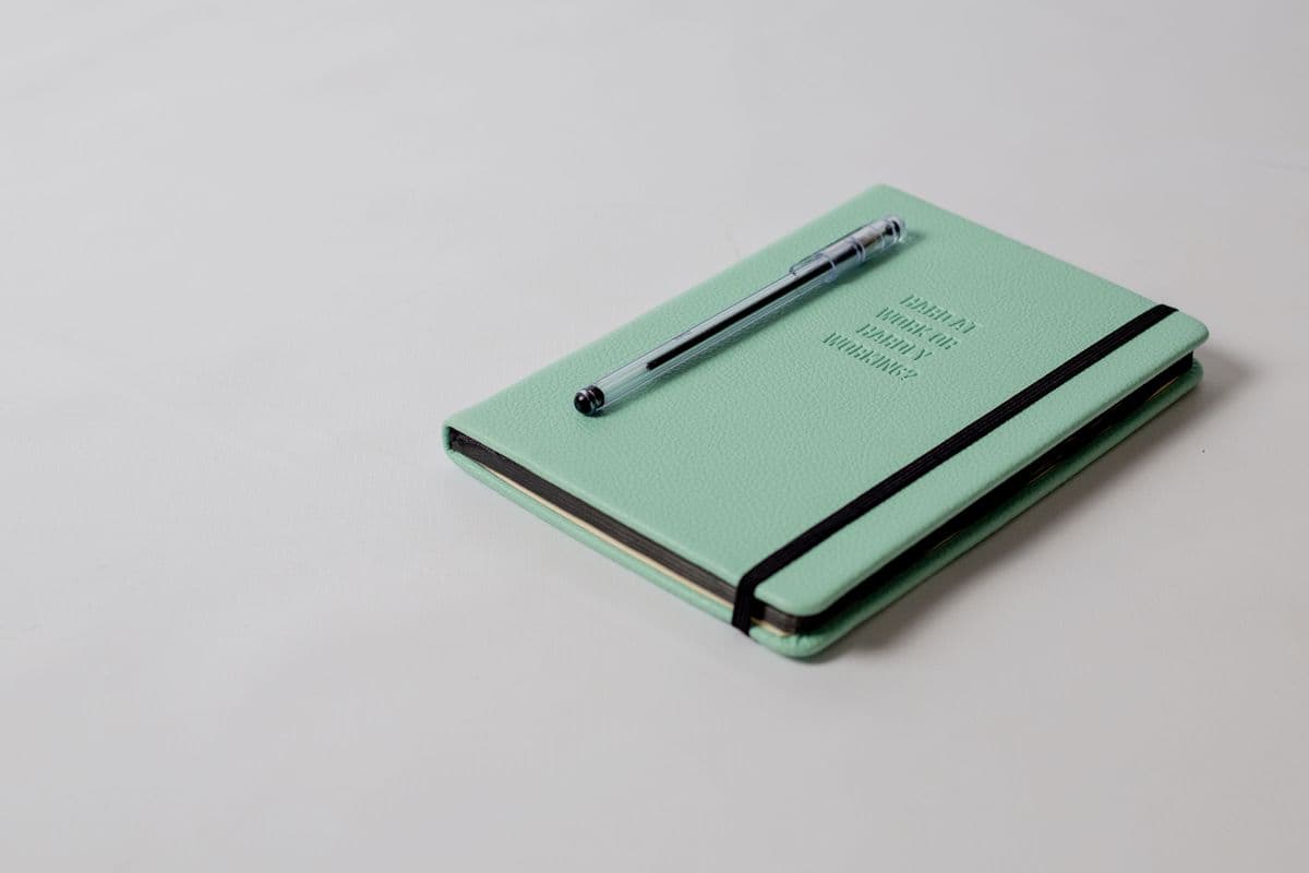 Buku agenda berwarna hijau toska dengan penutup elastis dan pena di atas meja.
