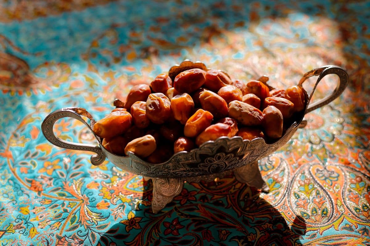 Semangkuk kurma segar dalam wadah perak artistik di atas kain bermotif cerah, oleh-oleh khas umroh yang wajib dibawa pulang.